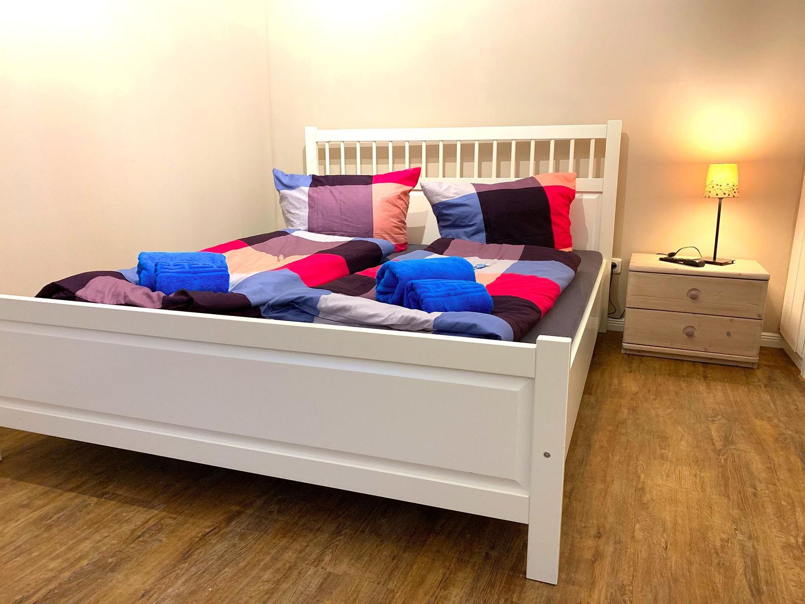 Schlafzimmer mit weißem Bett, buntem Bettzeug und Nachttisch mit Lampe.