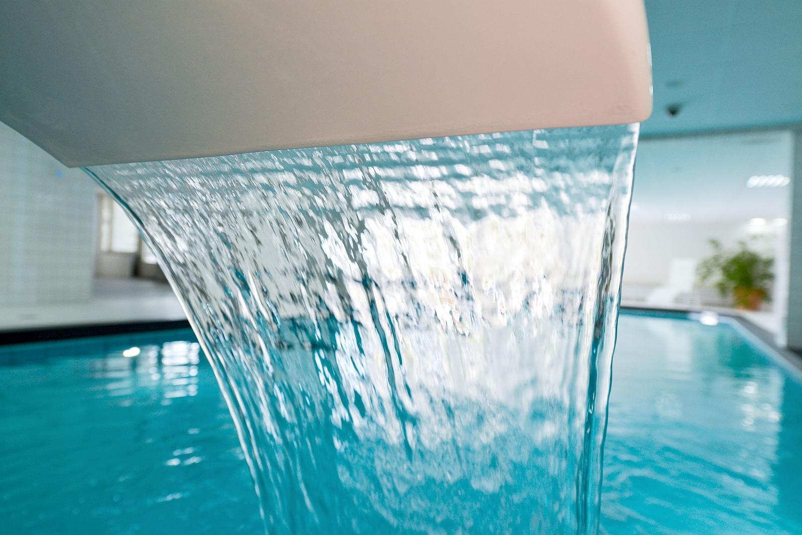 Wasser fällt aus einer Wand in den Pool. Im Hintergrund ist ein Raum mit Pflanzen.