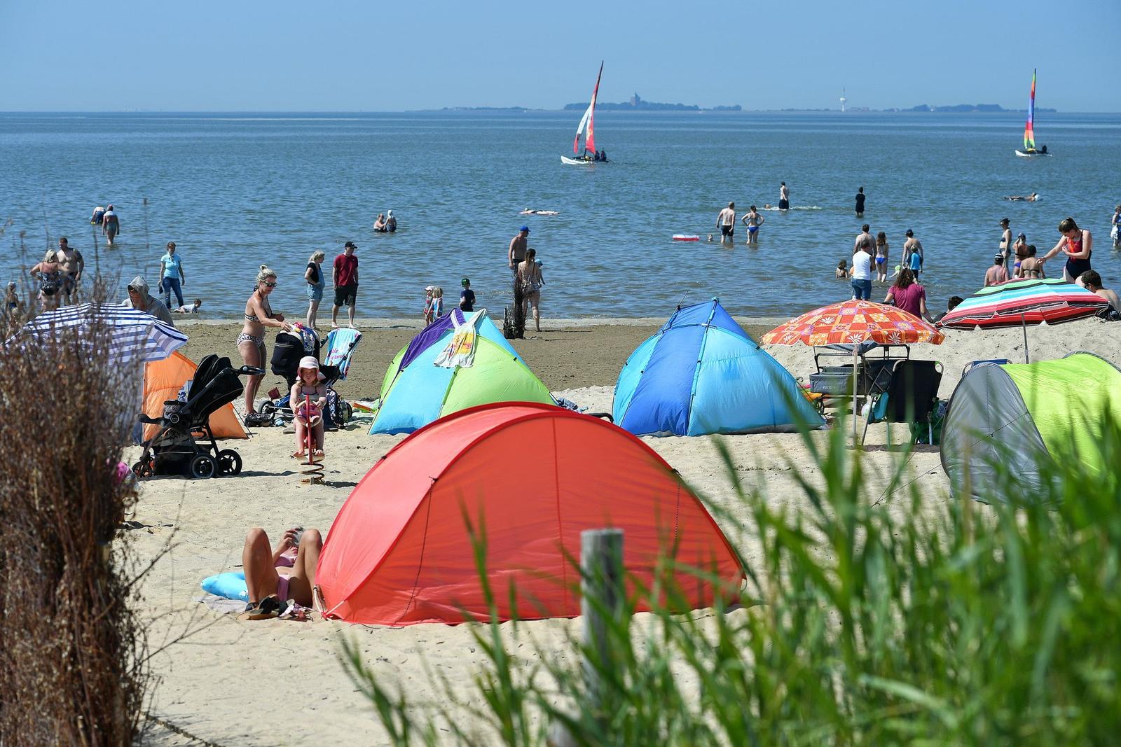 Strand mit bunten Zelten, Sonnenschirmen und Badegästen. Segelboote sind im Wasser.