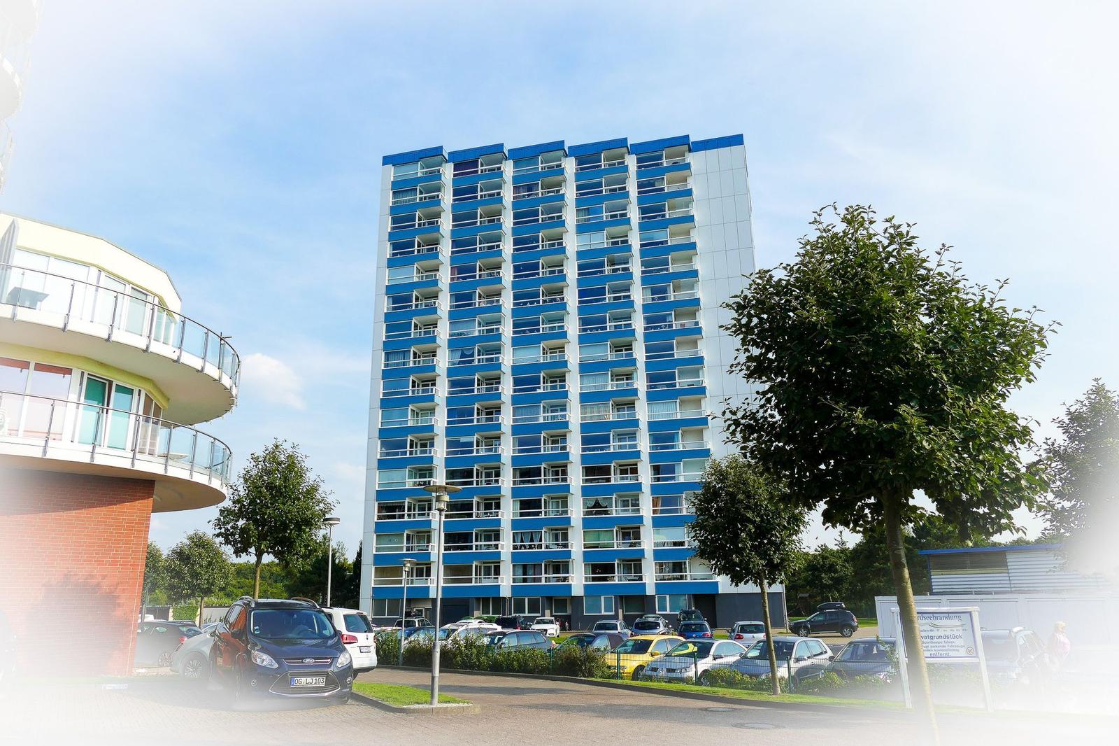 Hochhaus mit blauen Balkonen und Parkplatz vorne.