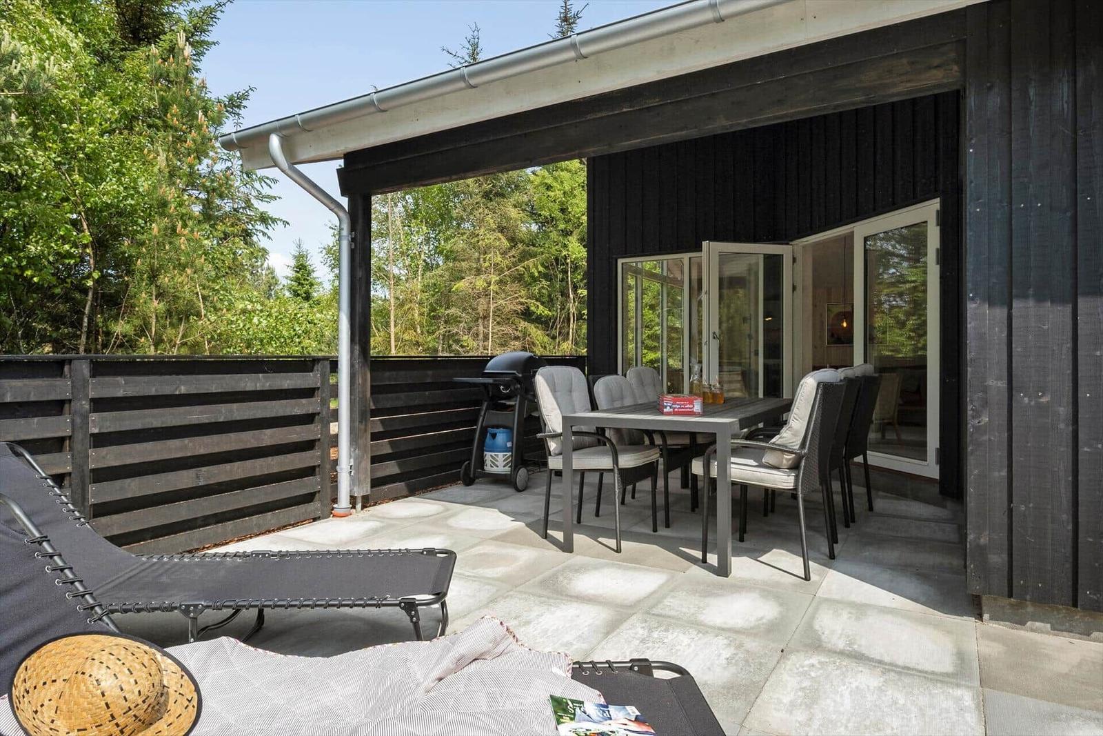 Terrasse mit Tisch, Stühlen, Grill und Liege unter schwarzem Holzverkleidung.