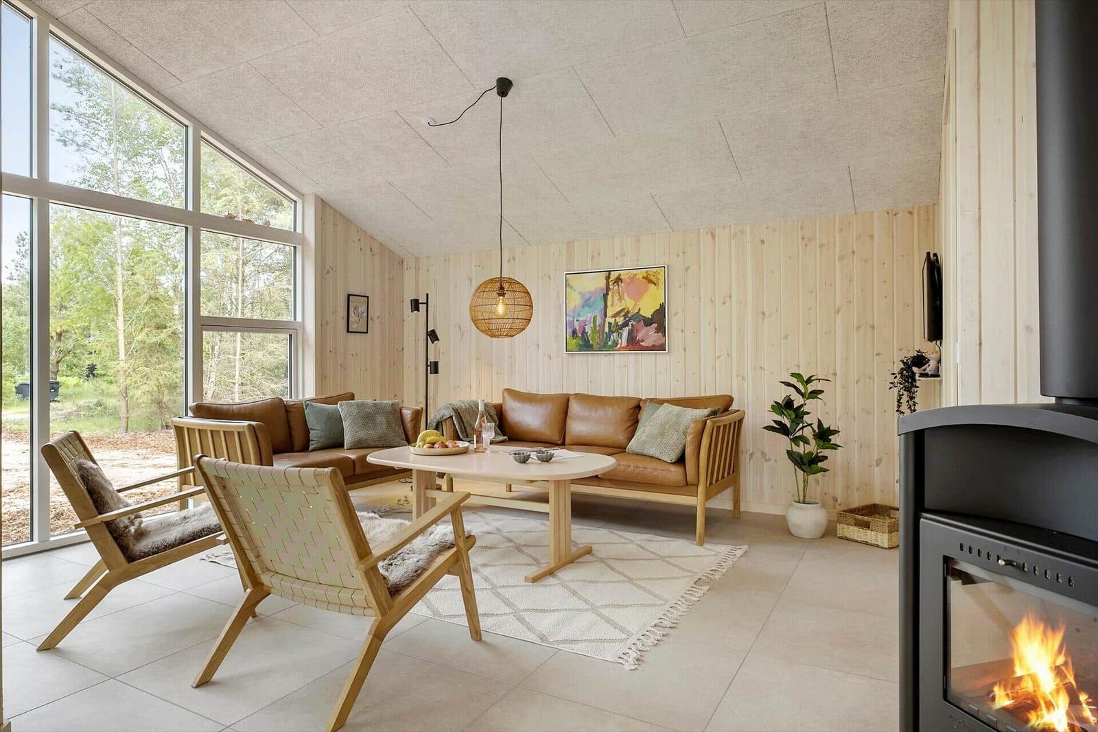 Wohnzimmer mit Holzwänden, Sofa, Sessel und Kaminofen. Große Fenster mit Blick auf Wald.