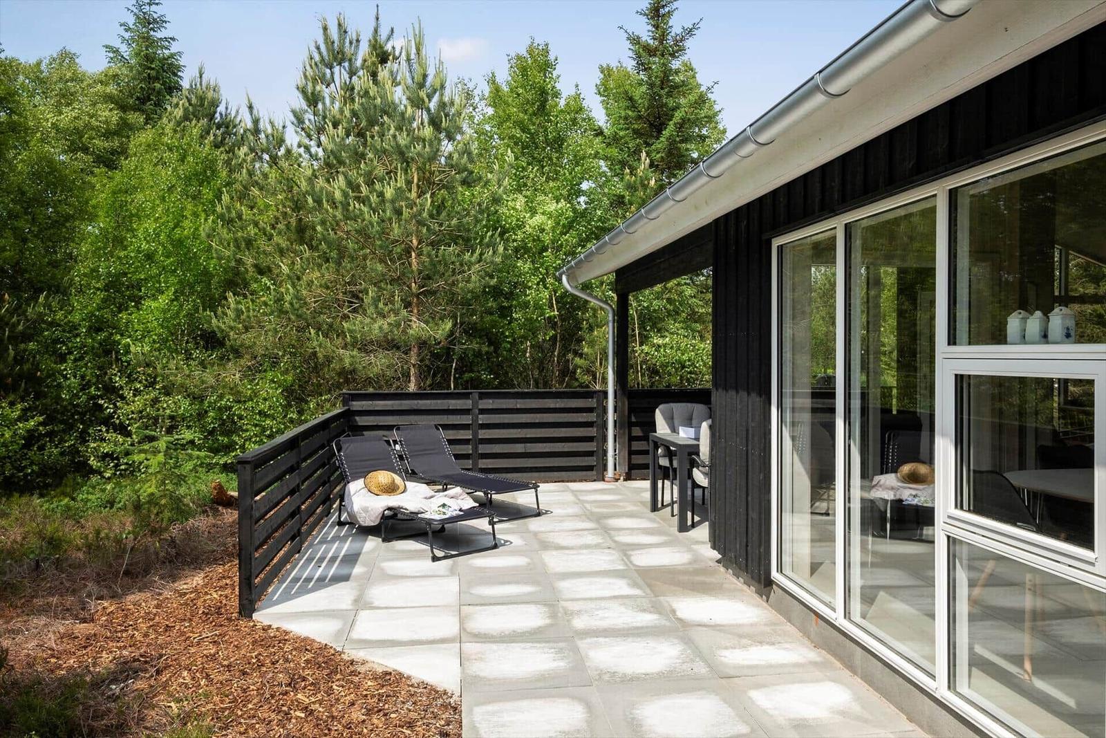 Terrasse mit Liegen und Tisch, umgeben von Wald und modernem Haus.