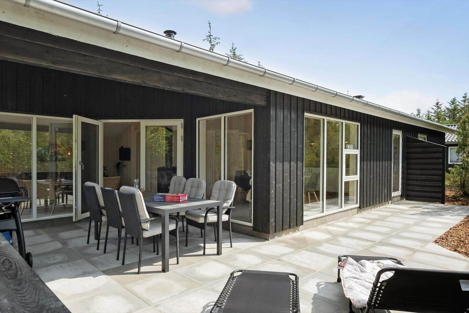 Terrasse mit Tisch, Stühlen und Liegestühlen vor modernem Haus mit großen Fenstern.