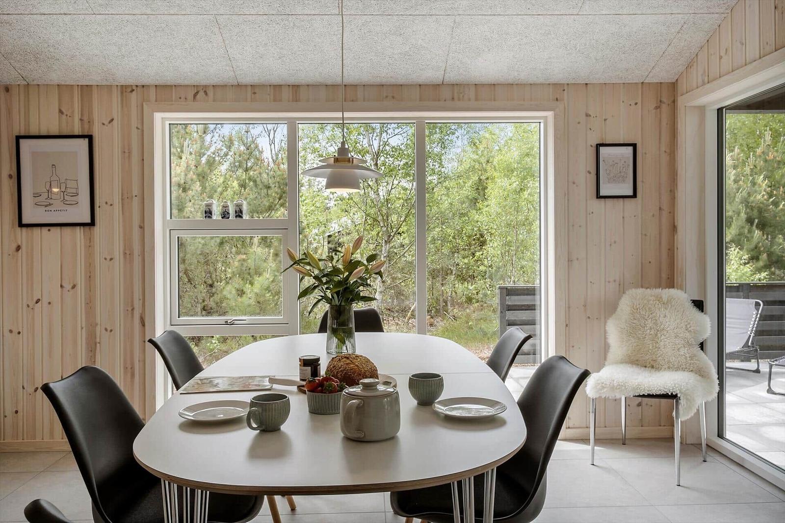 Esszimmer mit Holzwand, Tisch und Stühlen. Große Fenster zeigen Wald.