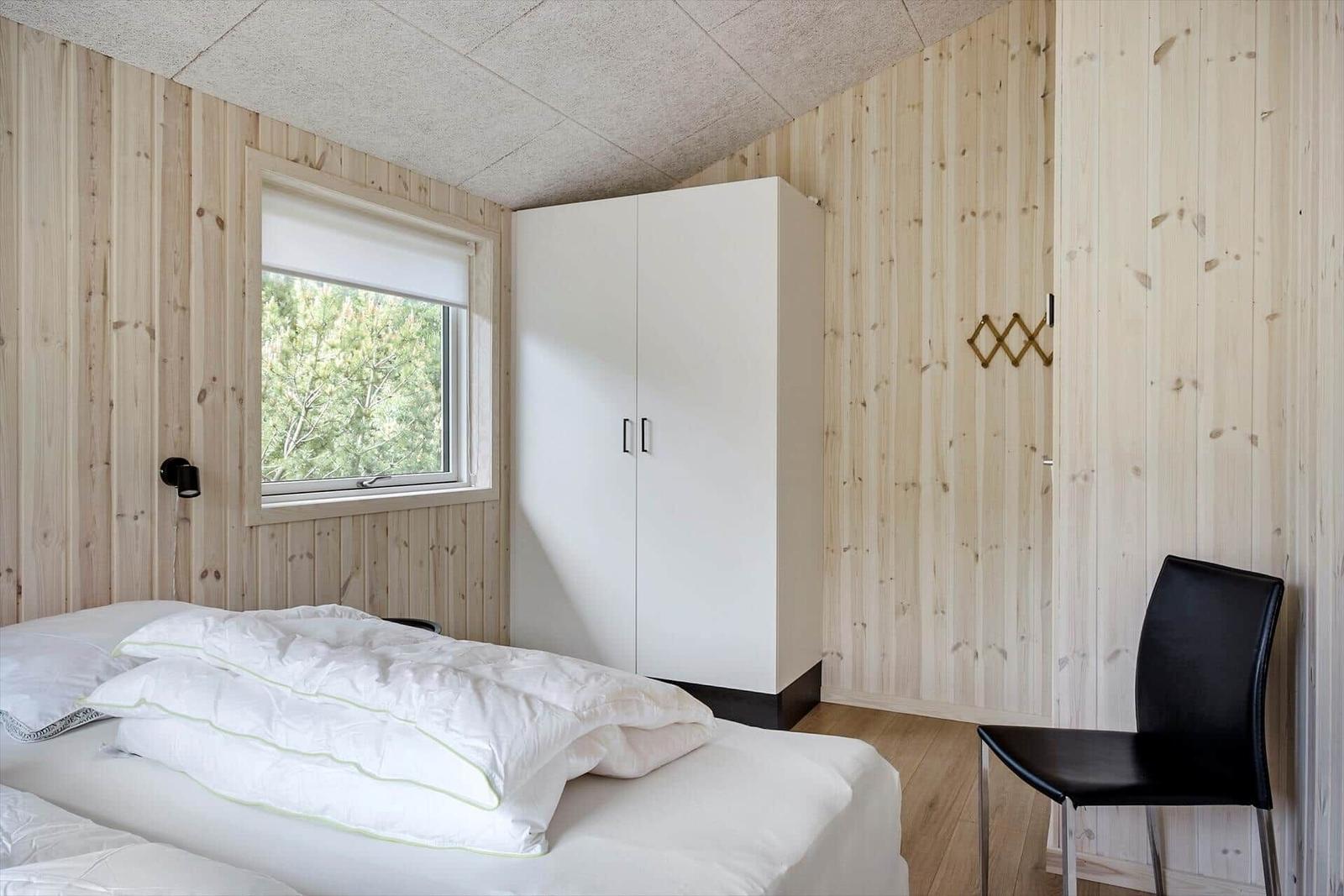Schlafzimmer mit Bett, Kleiderschrank und Stuhl. Holzwand und Fenster mit Blick auf Bäume.