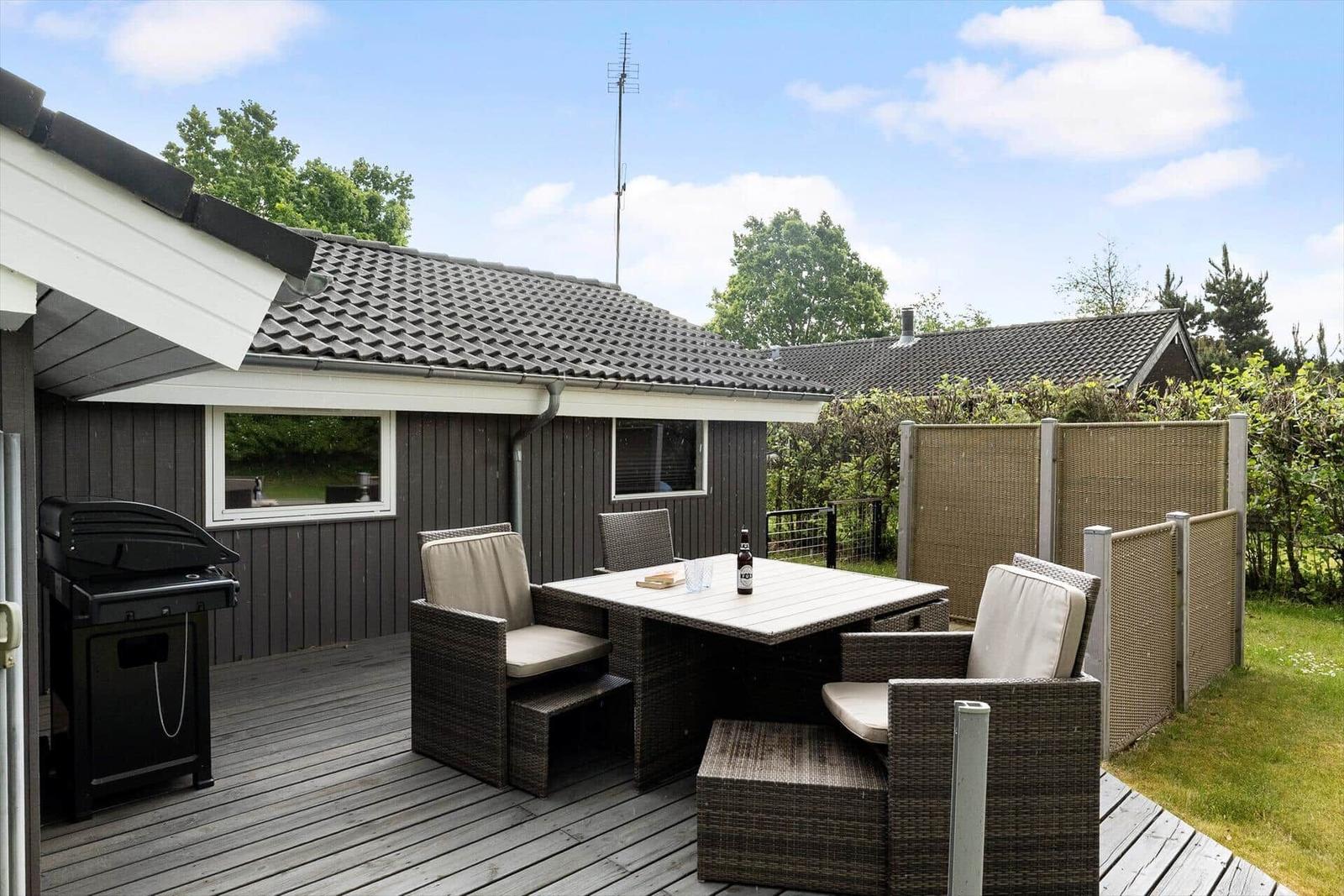 Terrasse mit Sitzgruppe, Tisch und Grill. Hintergrund: Holzhaus und Grünflächen.