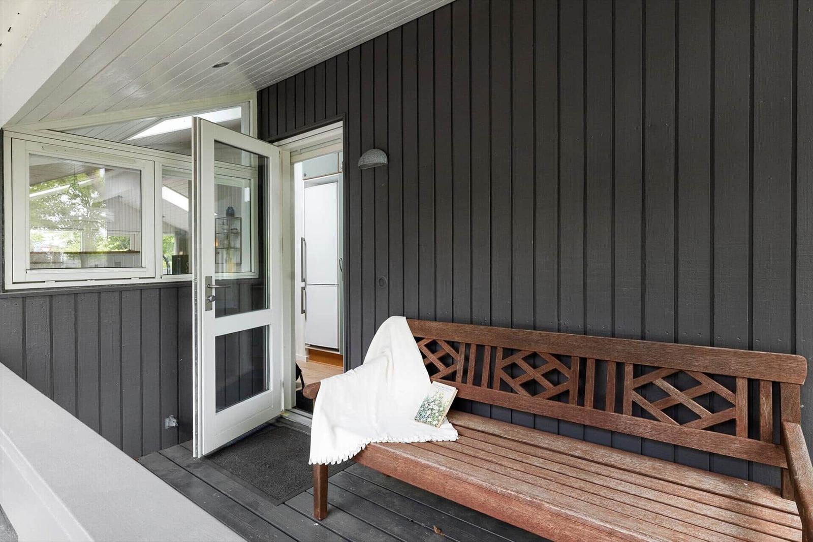 Holzbank mit weißer Decke und Buch auf Terrasse vor Haus mit dunkler Fassade.