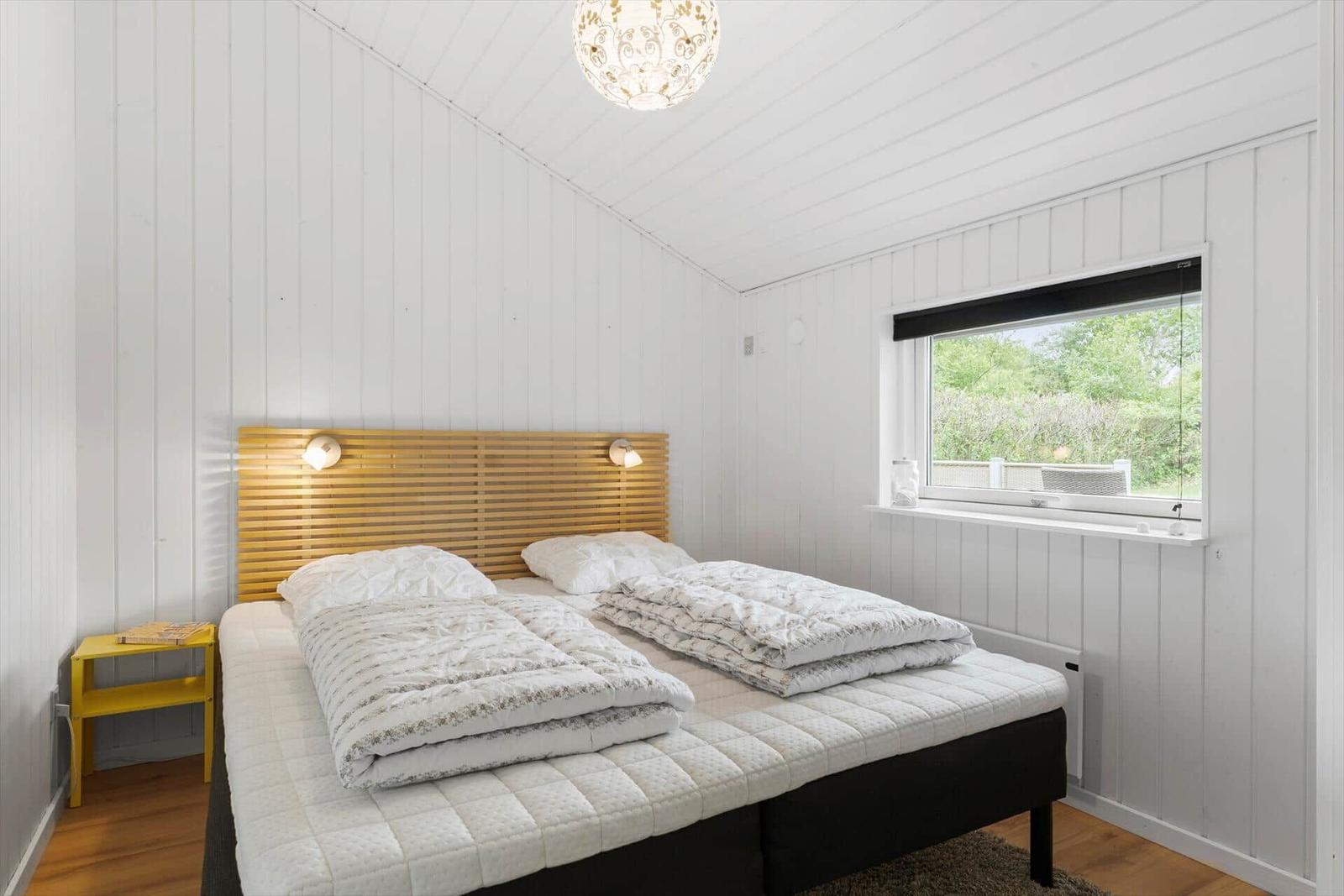 Schlafzimmer mit Bett, Holzwand und Fenster. Gelber Nachttisch und Decken.