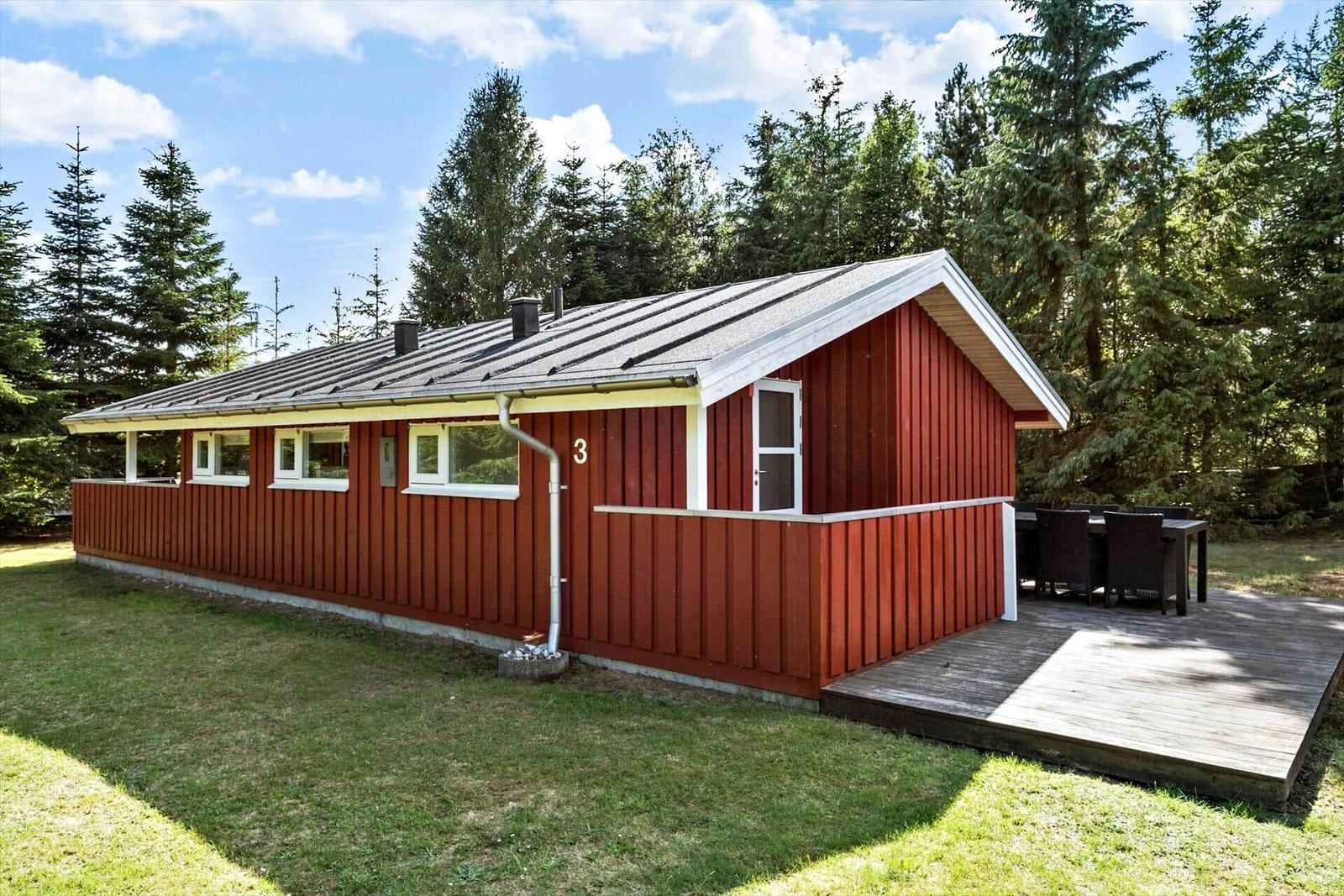 Rotes Holzhaus mit Terrasse und Garten. Umgeben von Bäumen unter blauem Himmel.