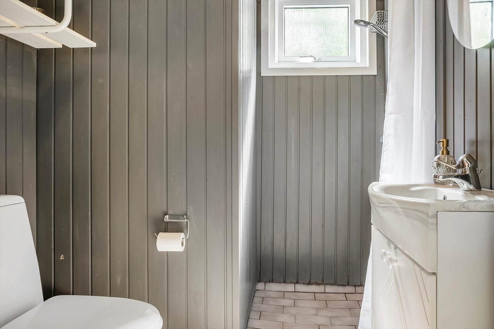 Badezimmer mit Toilette, Waschbecken und Dusche. Holzwand und Fenster sichtbar.