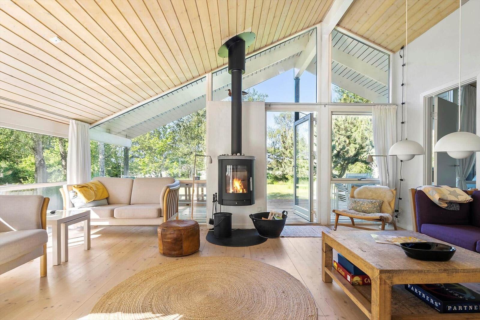 Wohnraum mit Kamin, Sofa und Holzboden. Große Fenster mit Blick auf Garten.