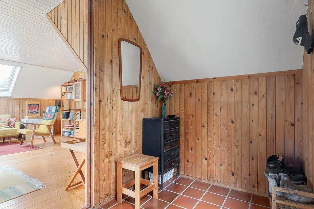 Holzwand, Spiegel, Schrank und Stuhl im Eingangsbereich. Im Hintergrund Wohnzimmer mit Sitzgelegenheit.