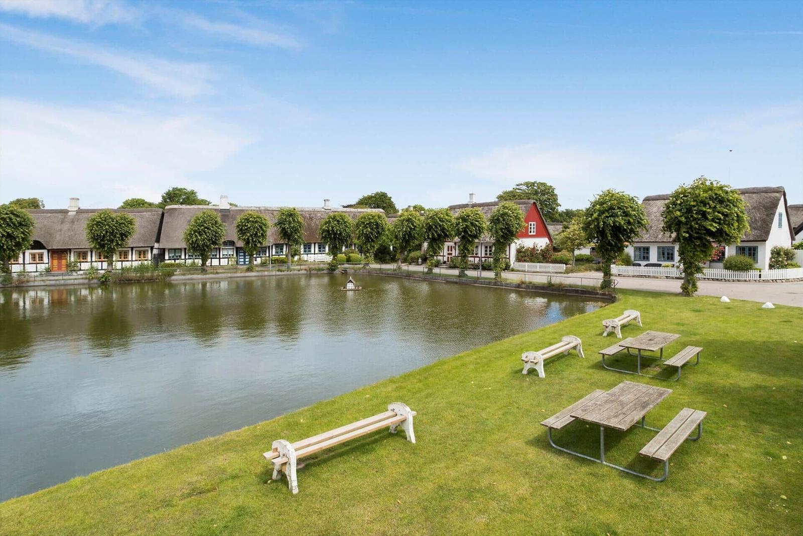 Grüner Rasenbereich mit Picknicktischen und Bänken am See. Dahinter stehen traditionelle Häuser mit Strohdächern.