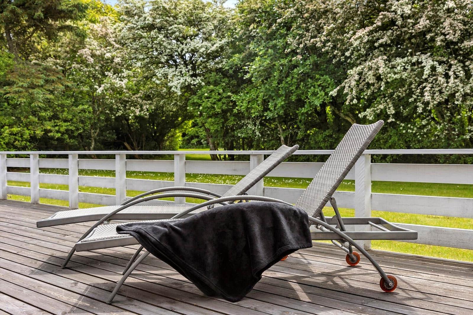 Two lounge chairs with dark towel on wooden terrace overlooking green trees.