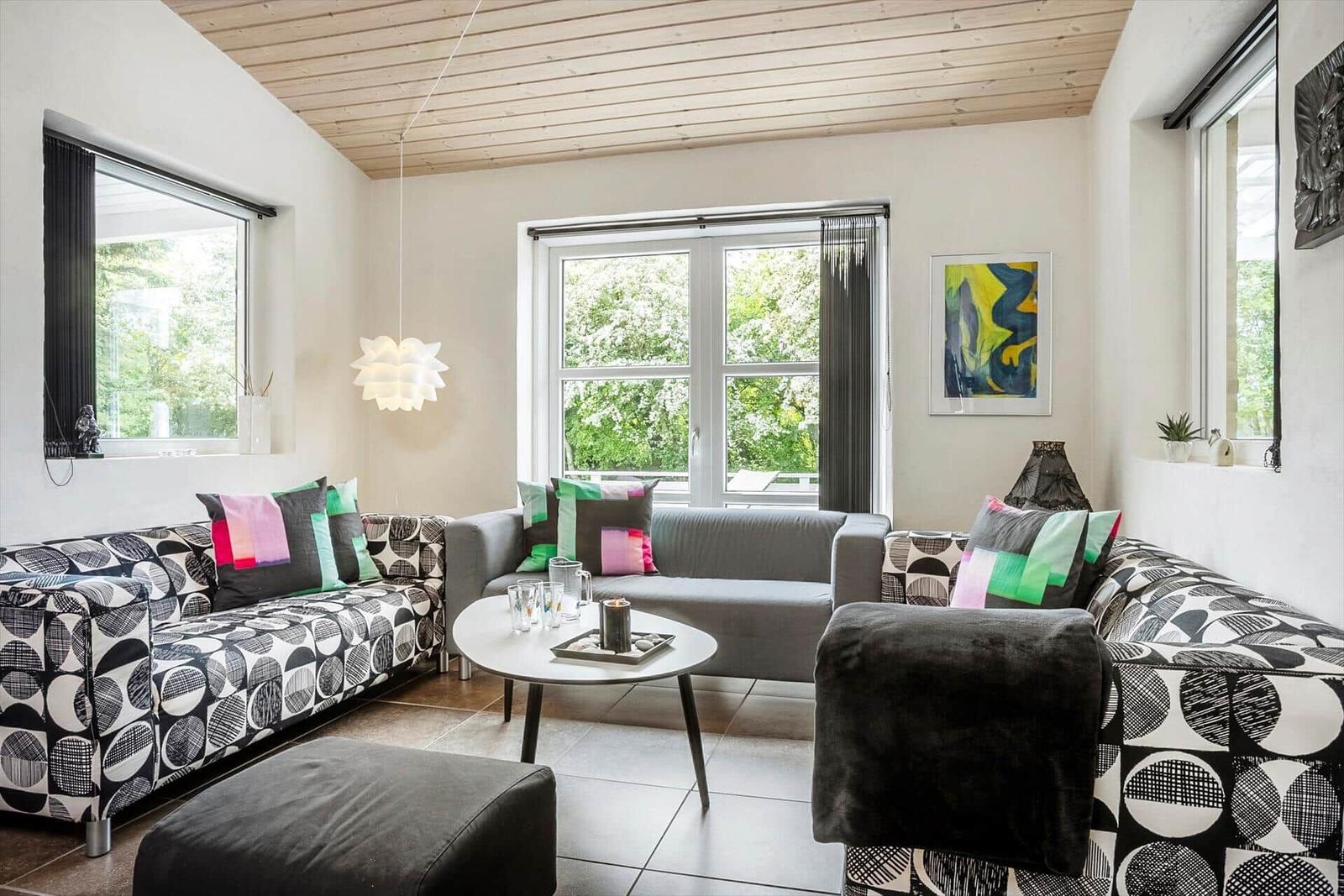 Living room with sofa, seating group, and table. Wooden ceiling and windows with view of green.