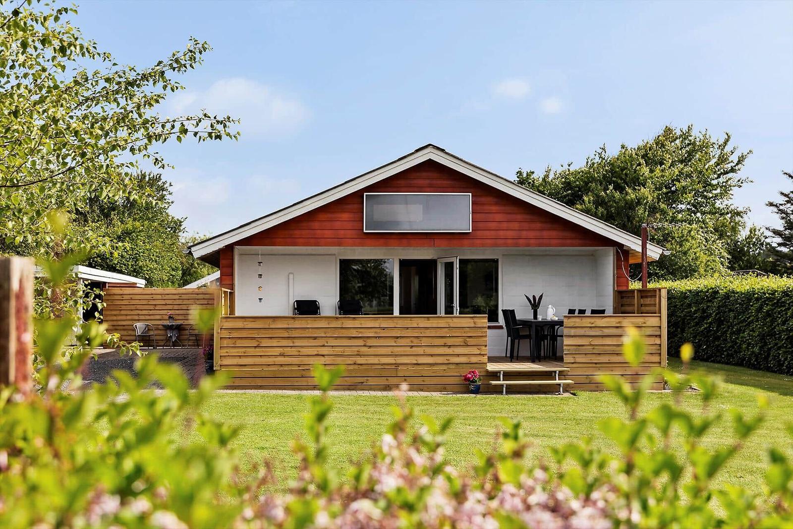 Red wooden house with terrace and garden. Black furniture and green trees surround the building.