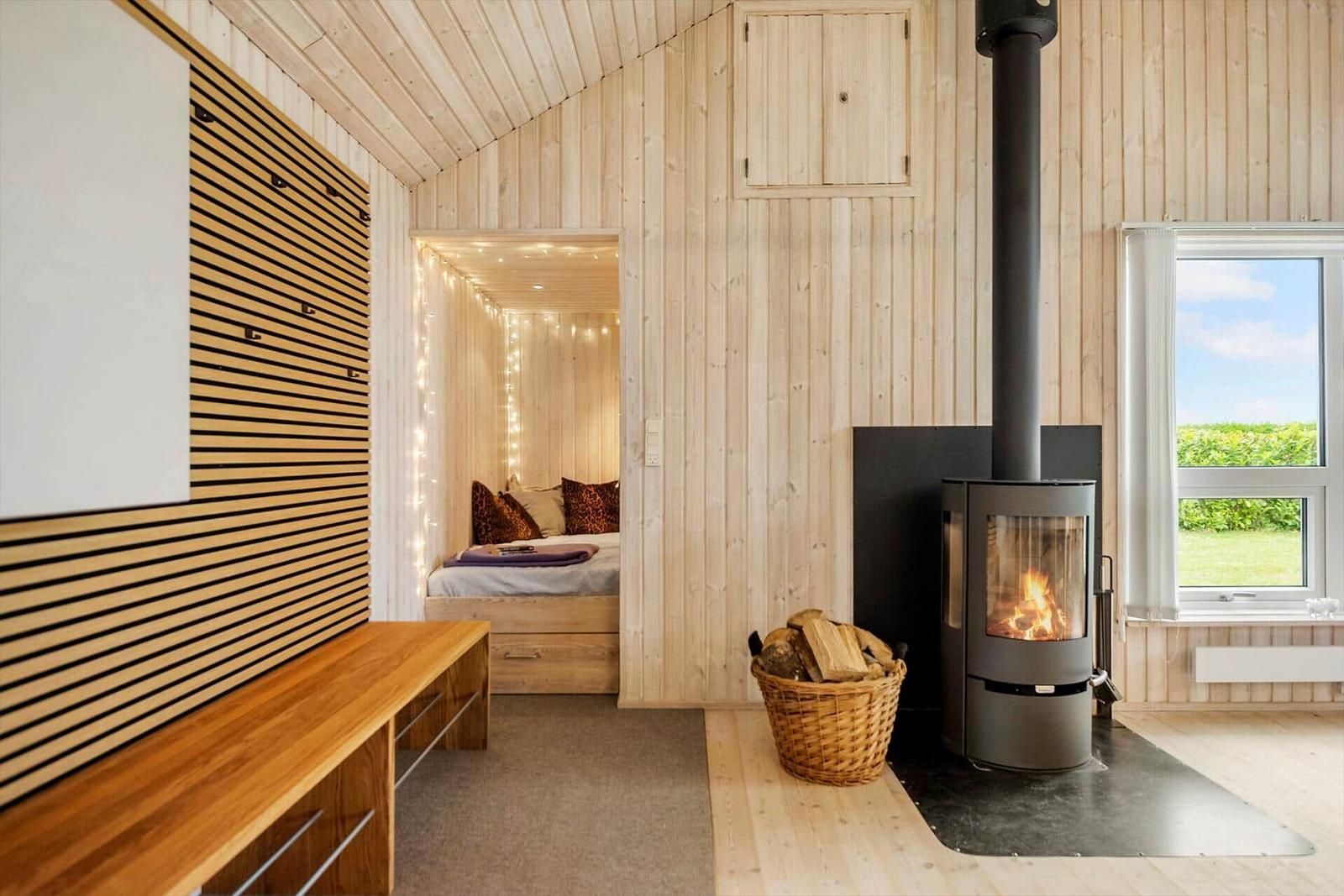 Wohnraum mit Kamin, Holzwand und Bett im Hintergrund. Fenster mit Blick auf Grünfläche.