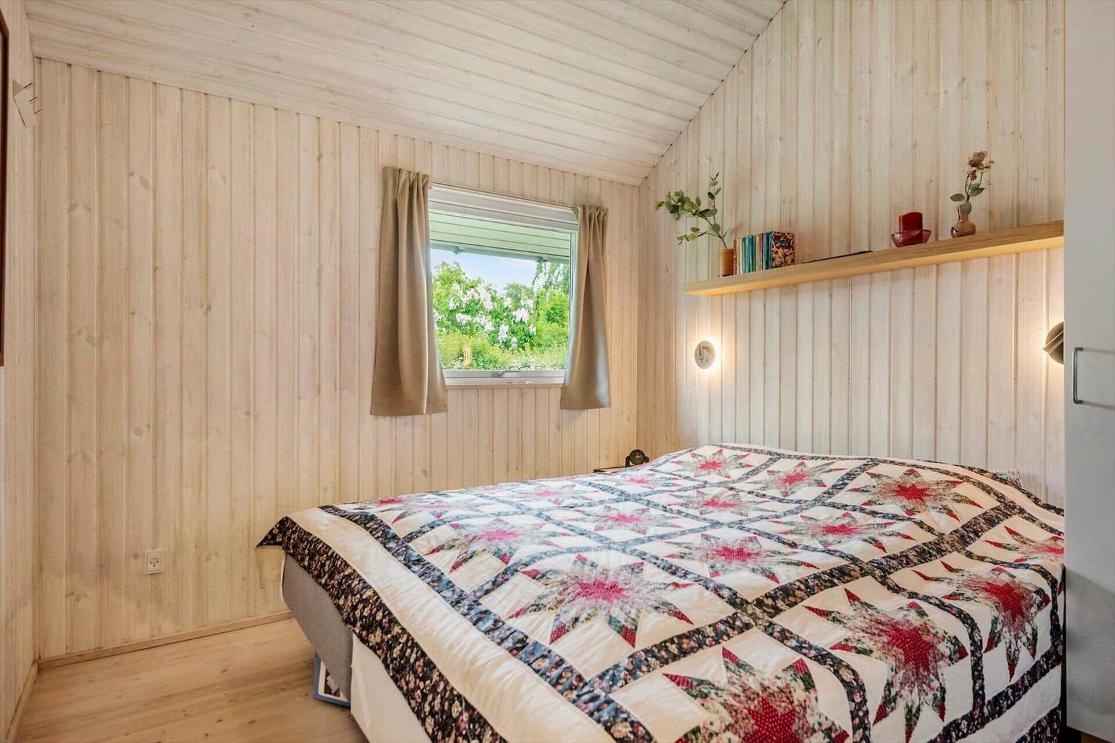 Schlafzimmer mit Holzwand, Bett mit gemusterter Decke und Fenster mit Blick auf Grün.