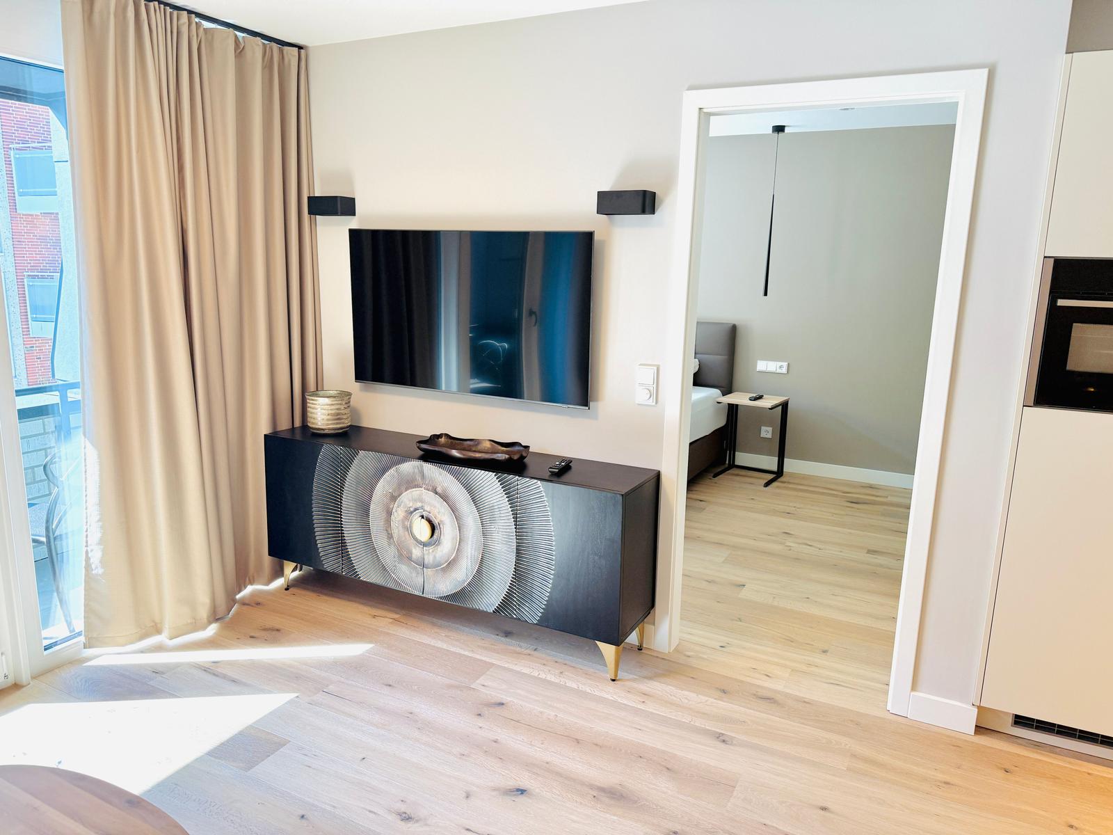 Wohnbereich mit TV, Sideboard und Blick in Schlafzimmer. Holzboden und Fenster mit Vorhängen.