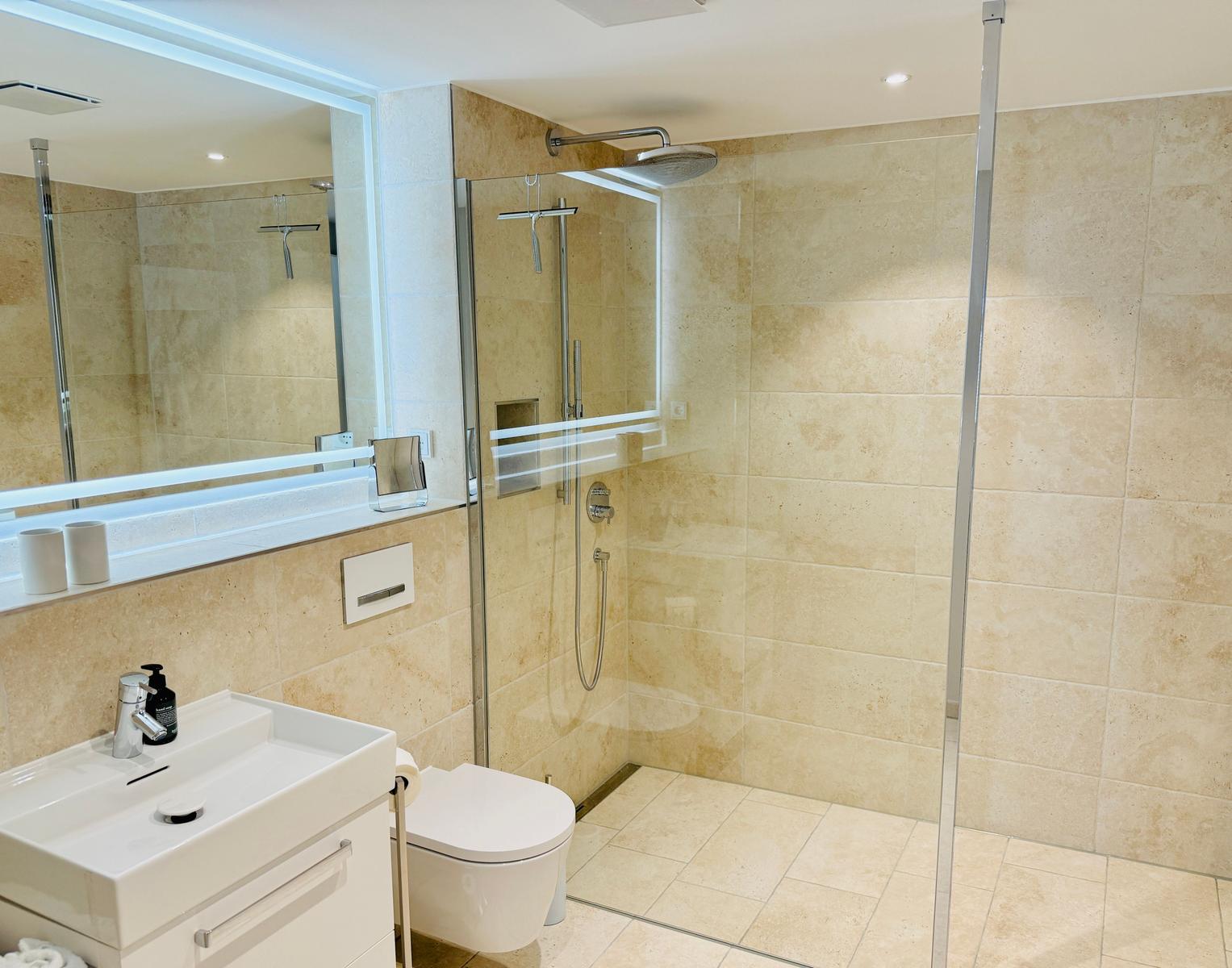 Badezimmer mit Dusche, Waschbecken und Toilette. Beleuchtetes Spiegel und Steinfliesen.