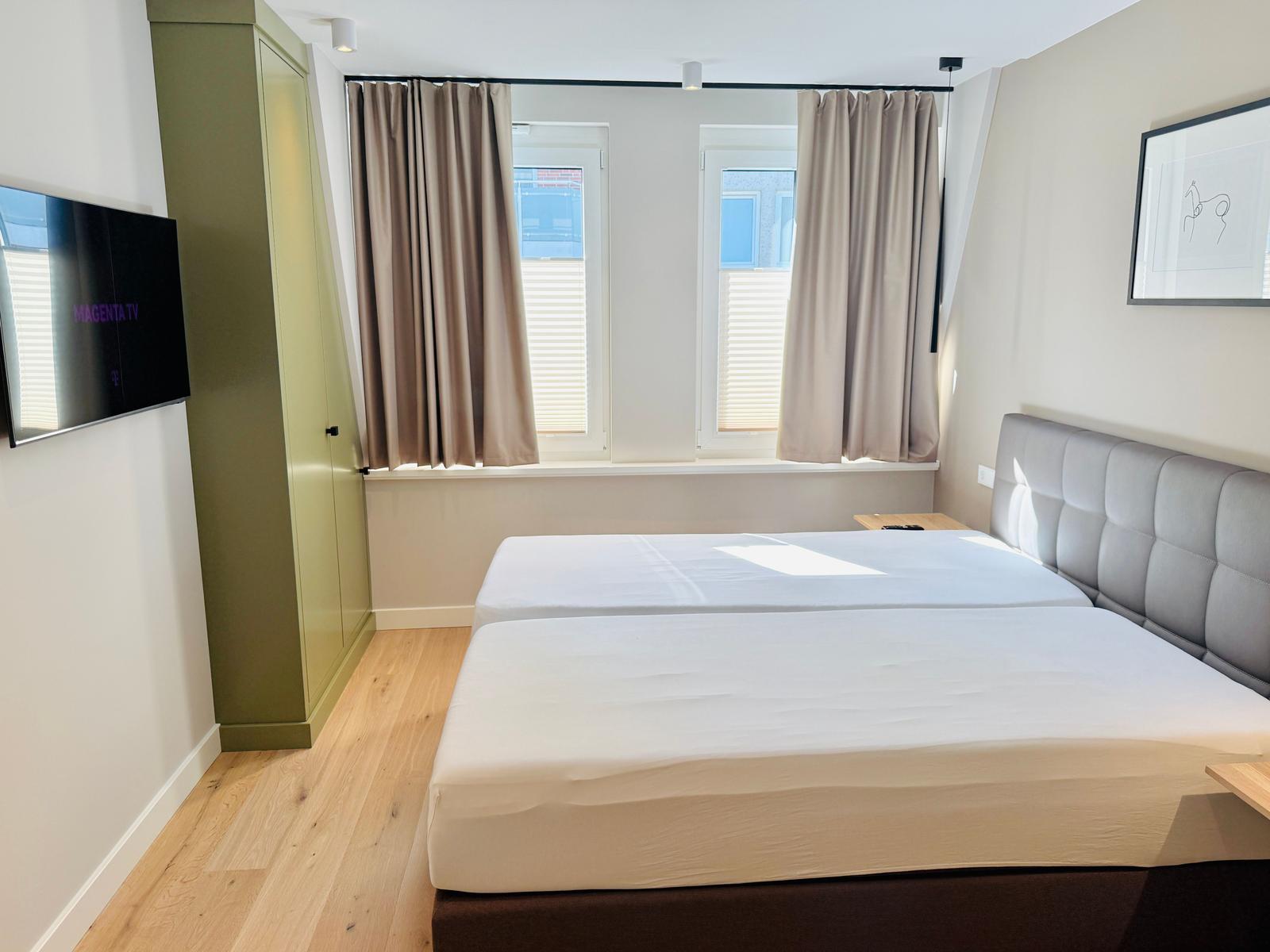 Schlafzimmer mit Doppelbett, Fenstern mit Vorhängen und Wand-TV.