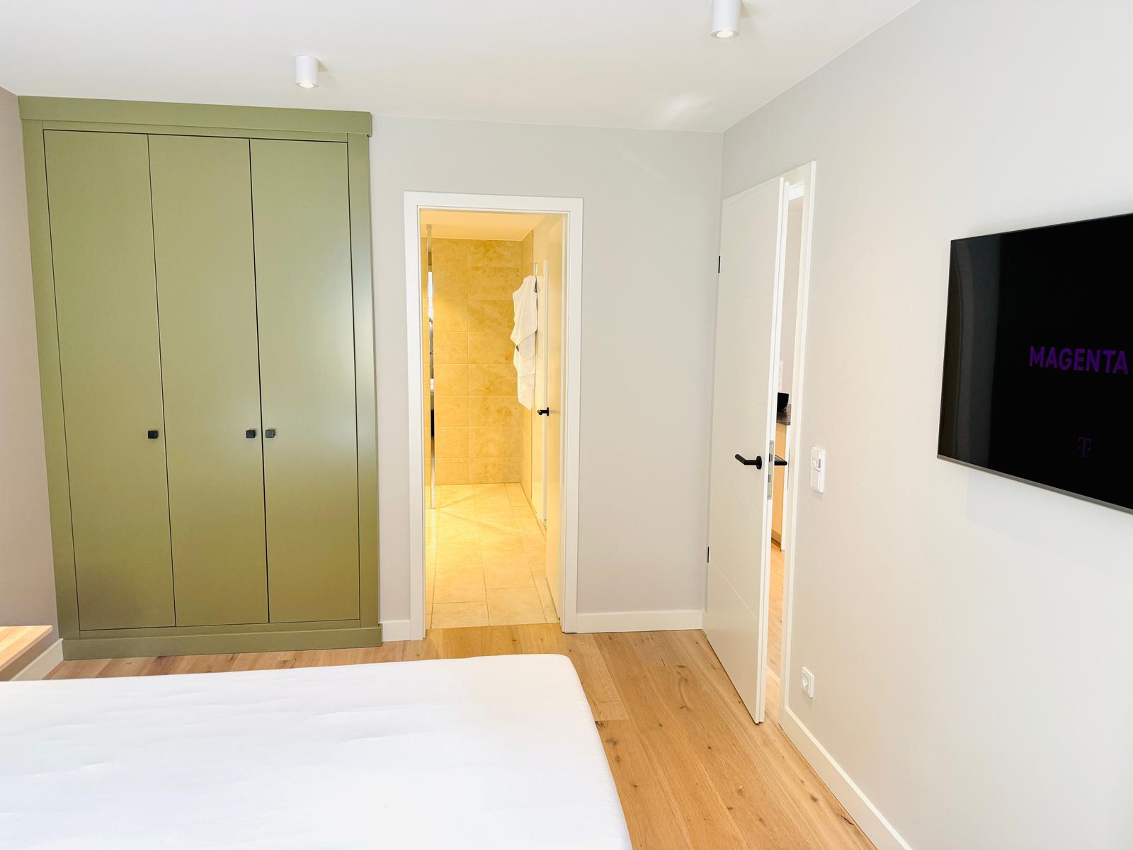 Schlafzimmer mit Bett, Kleiderschrank und TV. Tür führt in Bad.