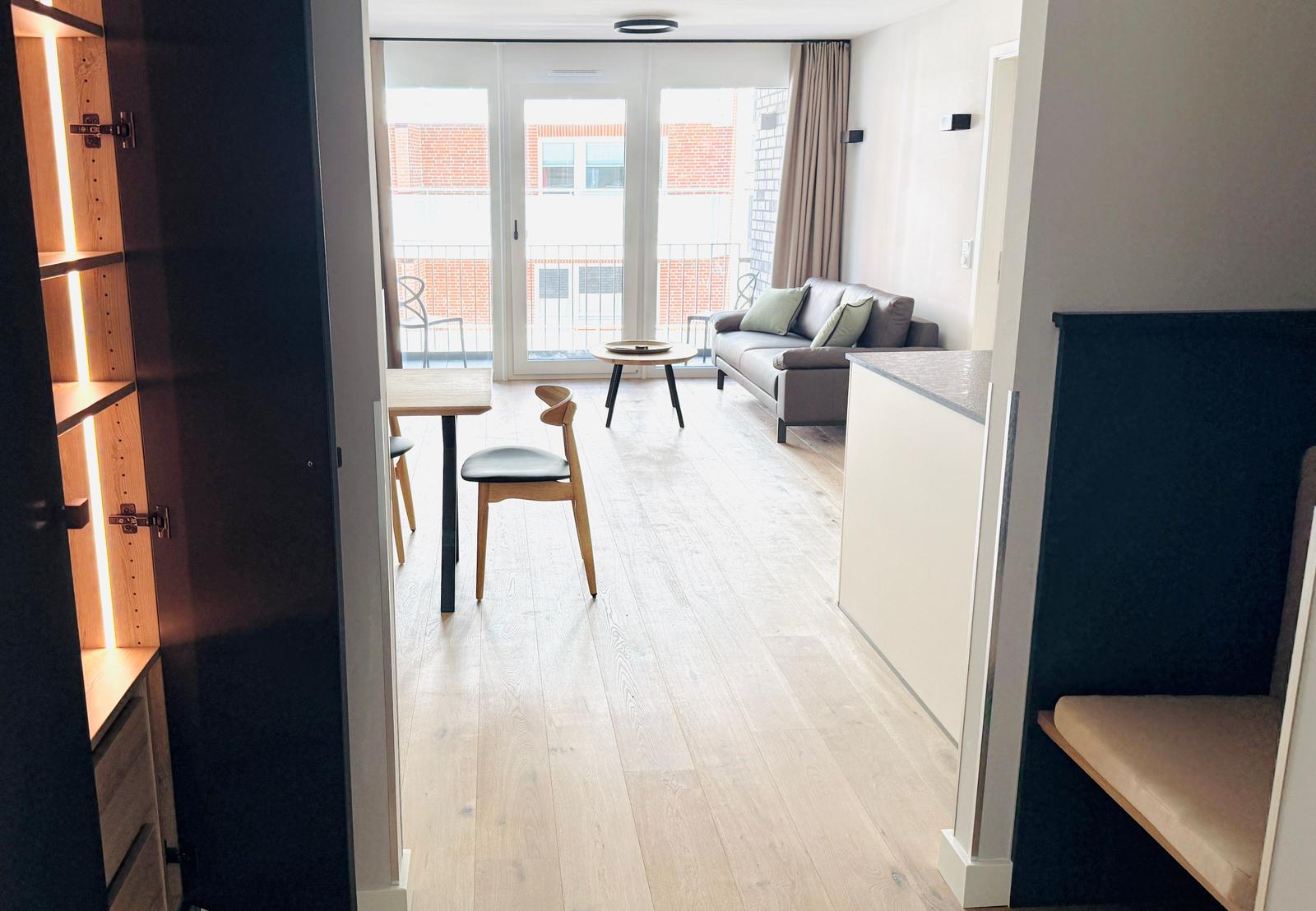 Wohnbereich mit Sofa, Tisch und Stühlen. Balkon mit Sitzgelegenheit. Holzboden und Küchenzeile.