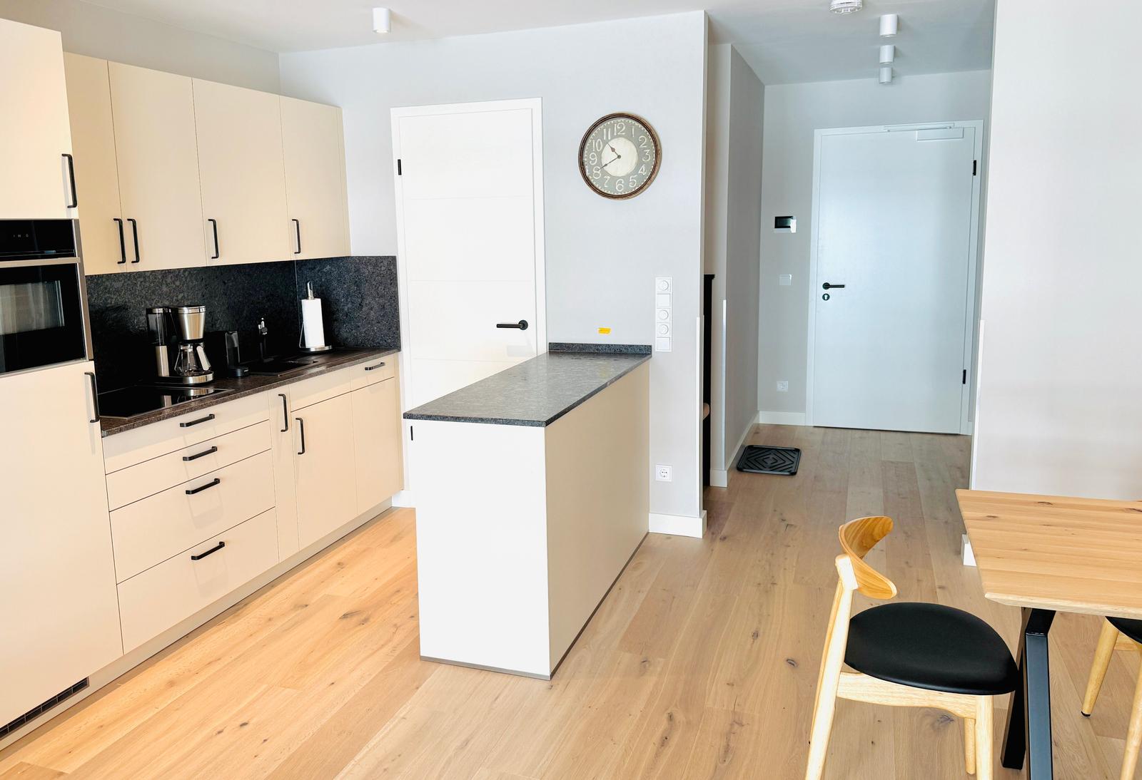 Küche mit Arbeitsplatte, Tisch und Stühle. Wanduhr und Holzboden sichtbar.