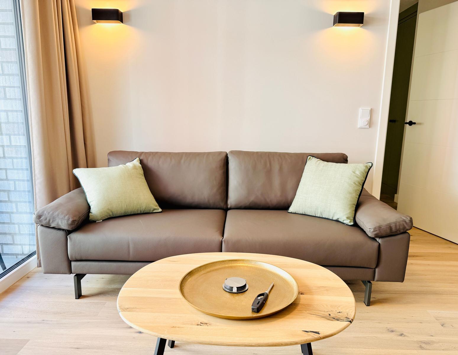 Ein braunes Sofa mit zwei Kissen steht vor einem Fenster. Davor ein runder Tisch mit einem goldenen Tablett.