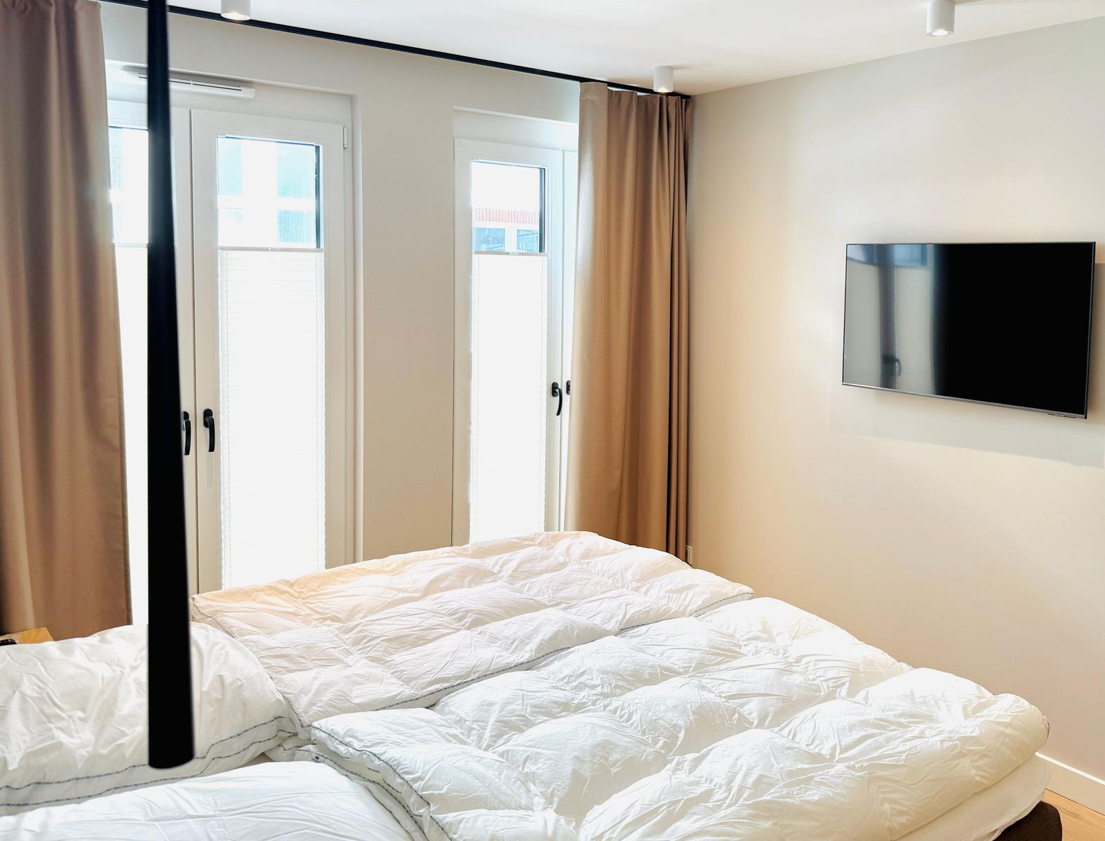 Schlafzimmer mit Bett, TV und Fenstern mit Vorhängen.