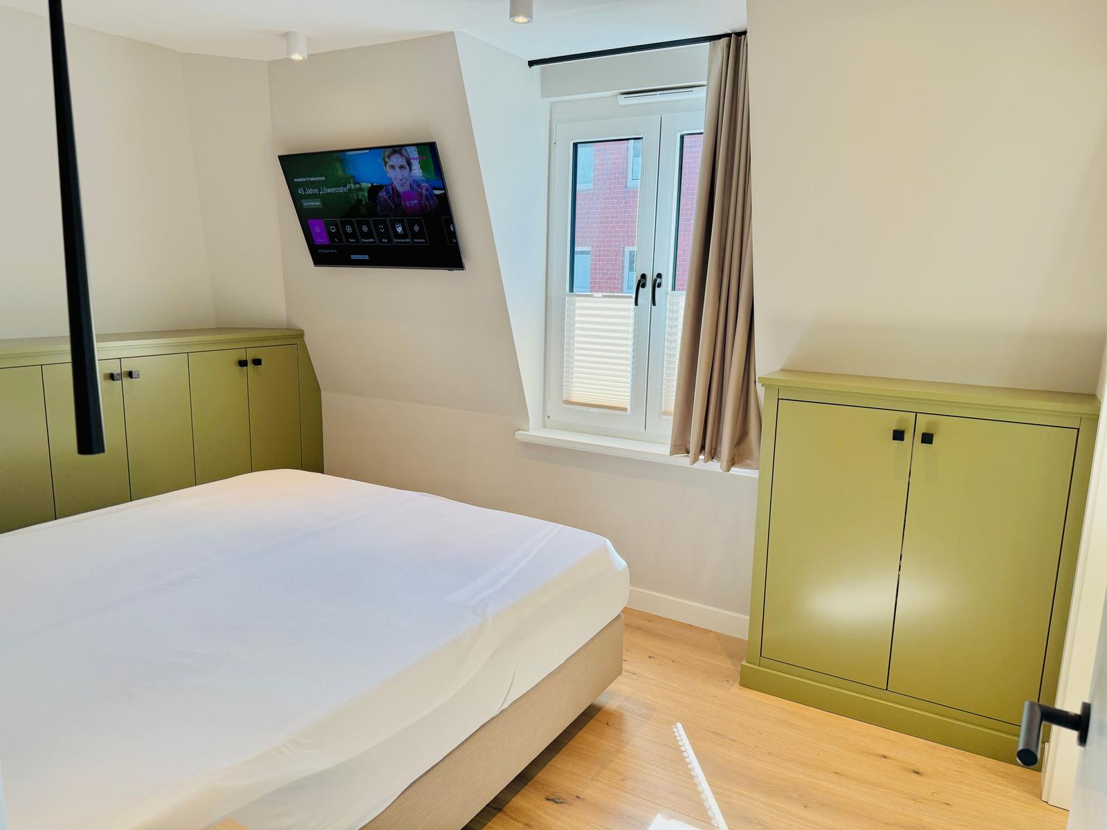 Schlafzimmer mit Bett, TV und Fenster. Grüne Schränke und Holzboden.