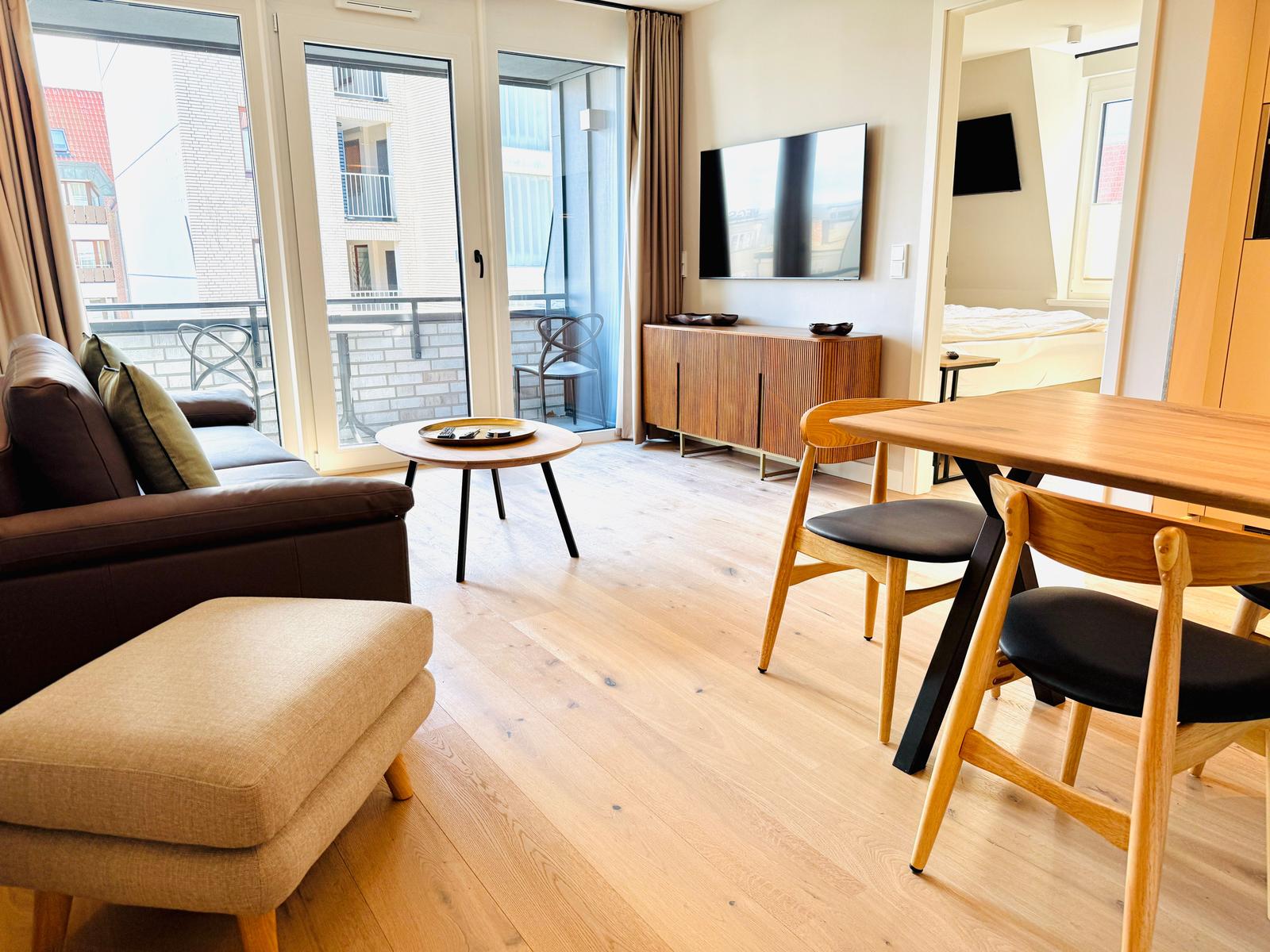 Wohn- und Essbereich mit Holzboden, Sofa, Esstisch und TV. Durchgang in Schlafzimmer.