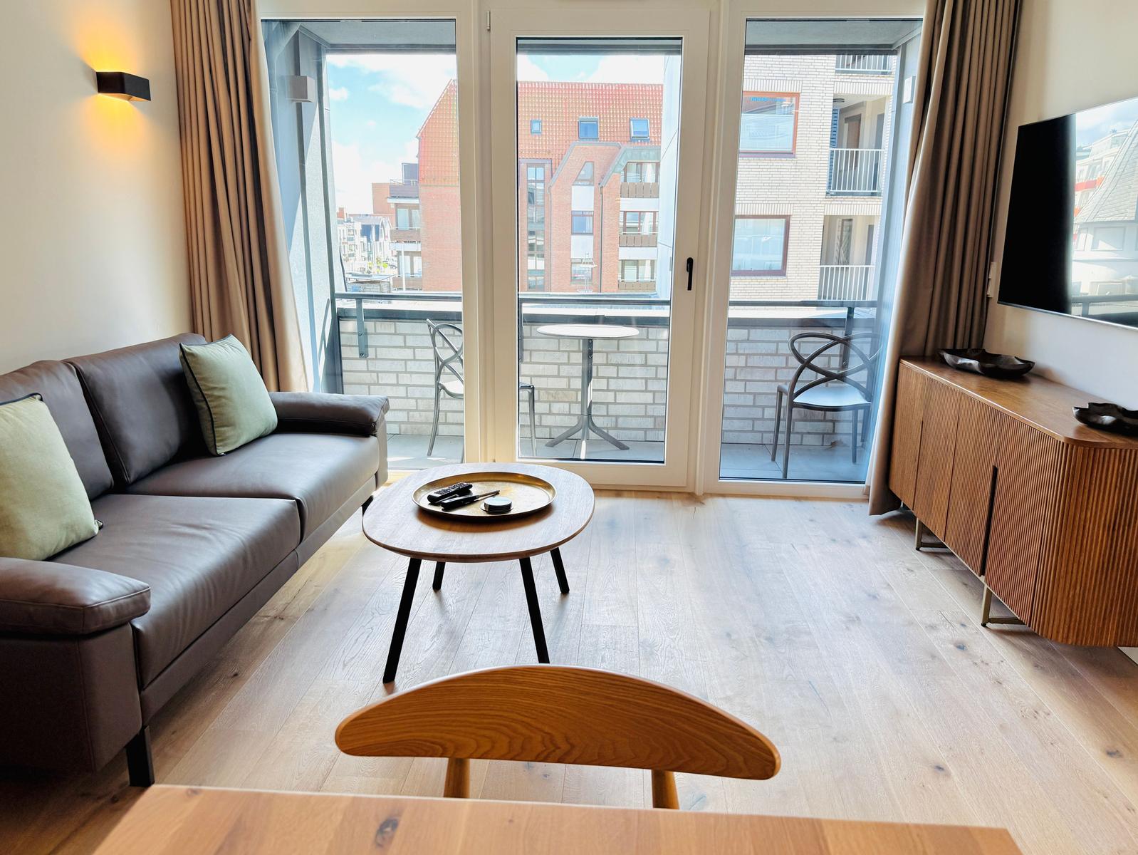 Wohnzimmer mit Sofa, Couchtisch und Balkon. Holzboden und Fernseher sichtbar.