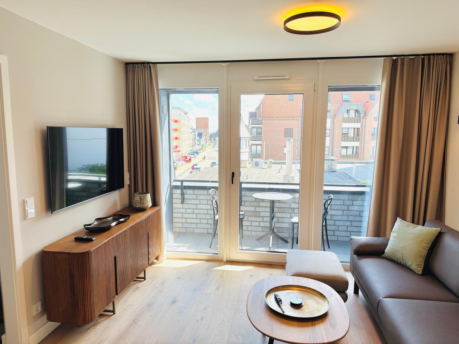 Wohnzimmer mit Sofa, Couchtisch und TV. Balkon mit Tisch und Stühlen. Blick auf Stadt.
