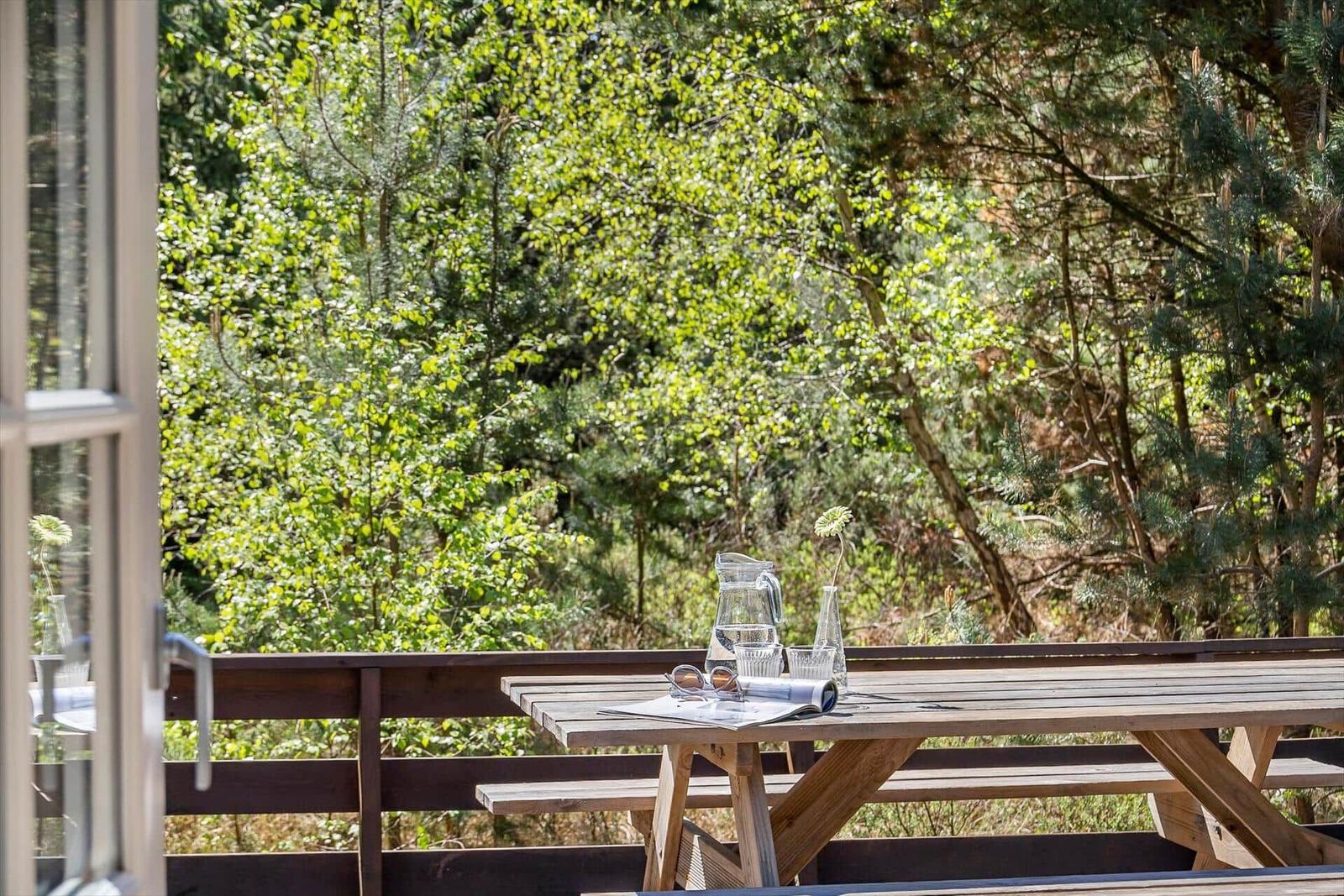Holztisch mit Wasserglas und Sonnenbrille auf Terrasse mit Waldblick.