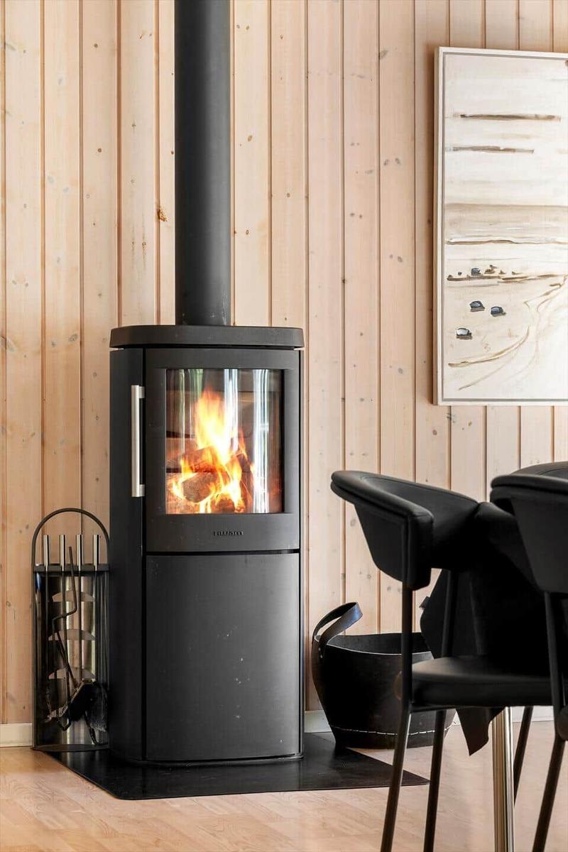Schwarzer Kaminofen mit brennendem Feuer in einem Raum mit Holzwandverkleidung und schwarzen Stühlen.