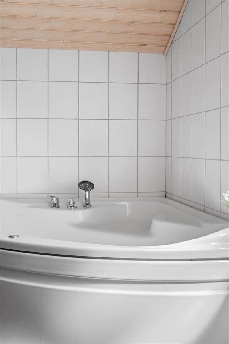 Duschkopf und Wasserhähne an einer weißen Badewanne mit weißen Fliesen und Holzdecke.