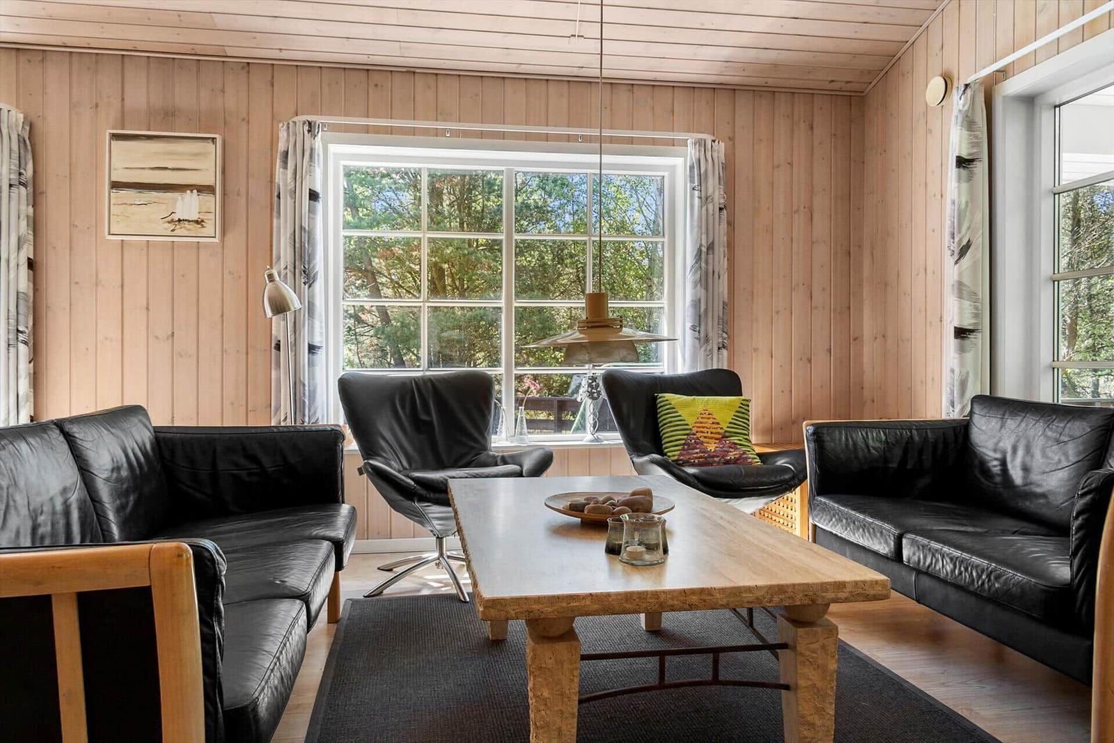 Wohnzimmer mit schwarzen Ledermöbeln, Holztisch und Holzwänden. Große Fenster mit Blick auf Bäume.