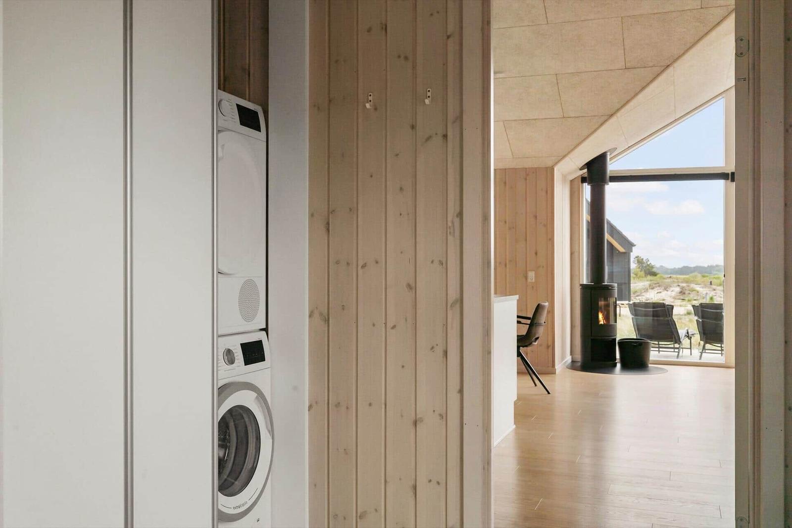 Wäschetrockner und Waschmaschine in einem Raum mit Holzwand. Durchgang in Wohnbereich mit Kamin und Fenster.