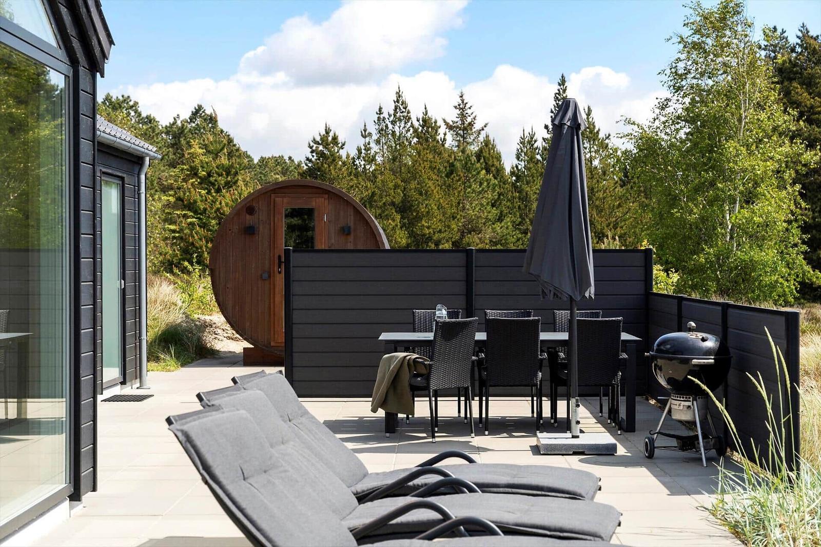 Terrasse mit Liegen, Tisch, Stühlen und Grill. Hintergrund: Wald und runder Holzaufbau.