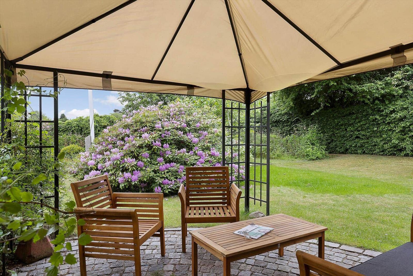 Terrace with sunshade, wooden chairs and table. Green lawn and flowering bushes in background.