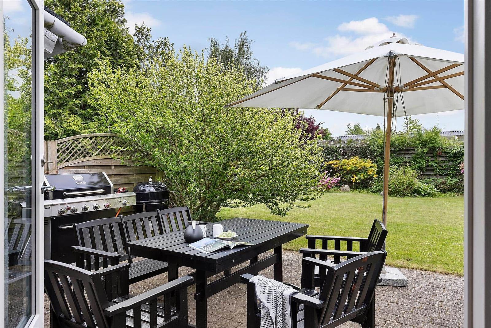 Terrace with table, chairs, and umbrella. Grill and lawn in the background.