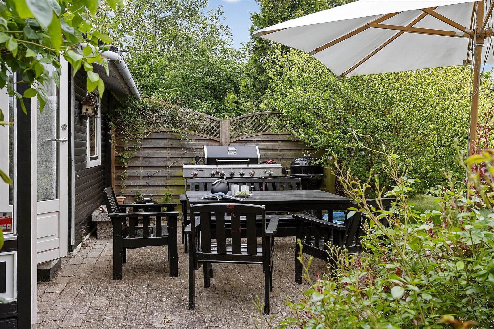 Terrace with table, chairs, grill, and umbrella. Background: wooden fence and greenery.