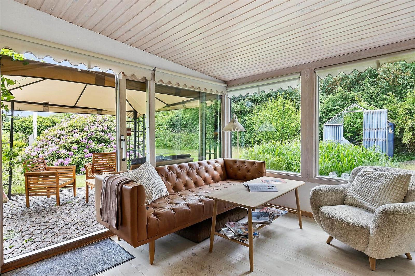Living area with brown leather sofa, wooden table, and armchair. Large windows show a garden.