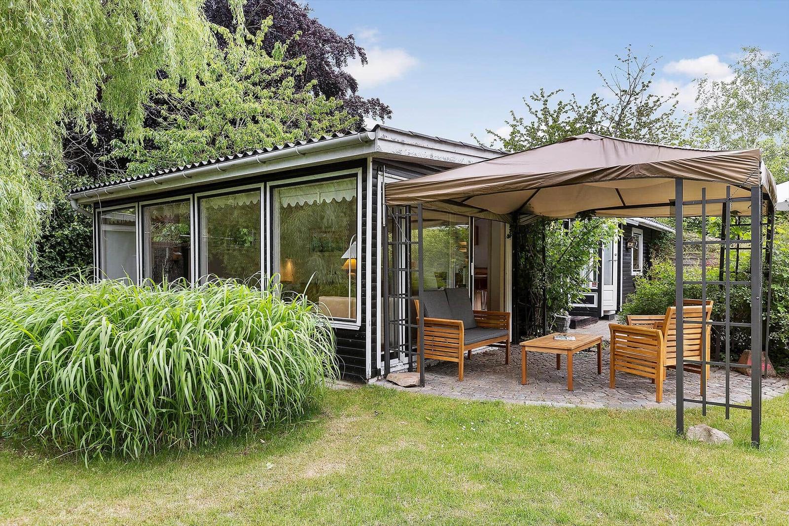 House with terrace, awning, and garden furniture. Green areas and trees surround the building.