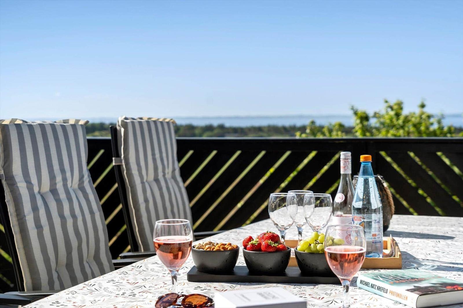 Terrace with table, chairs, and sea view. On the table are glasses, fruit, and bottles.