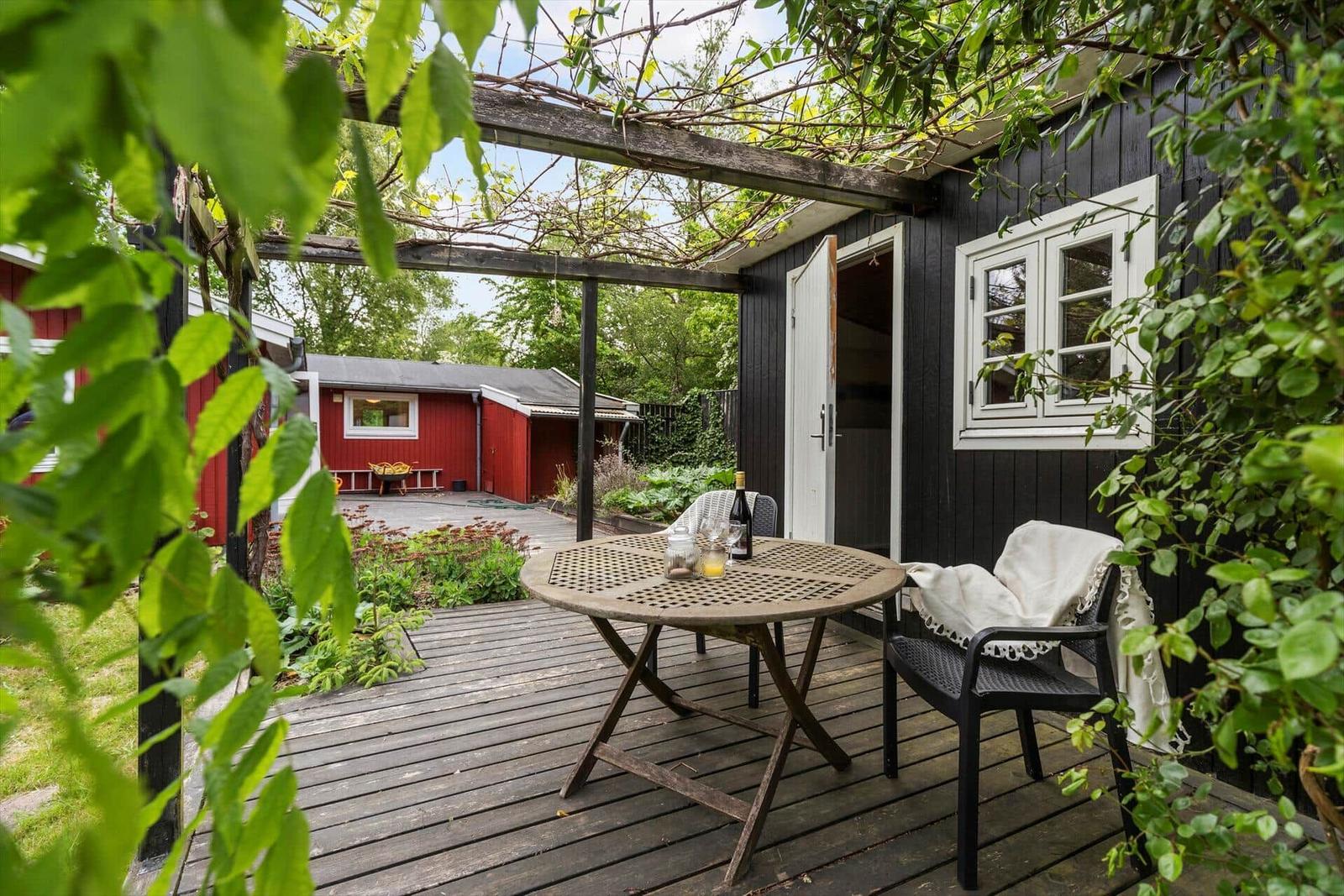 Terrasse mit Tisch und Stühlen unter Pergola. Schwarzes Haus mit weißem Fenster. Grün umgeben.