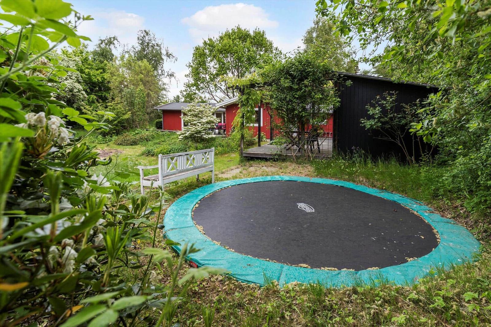 Ein Trampolin im Garten vor einem roten Haus mit weißem Gartenbank.