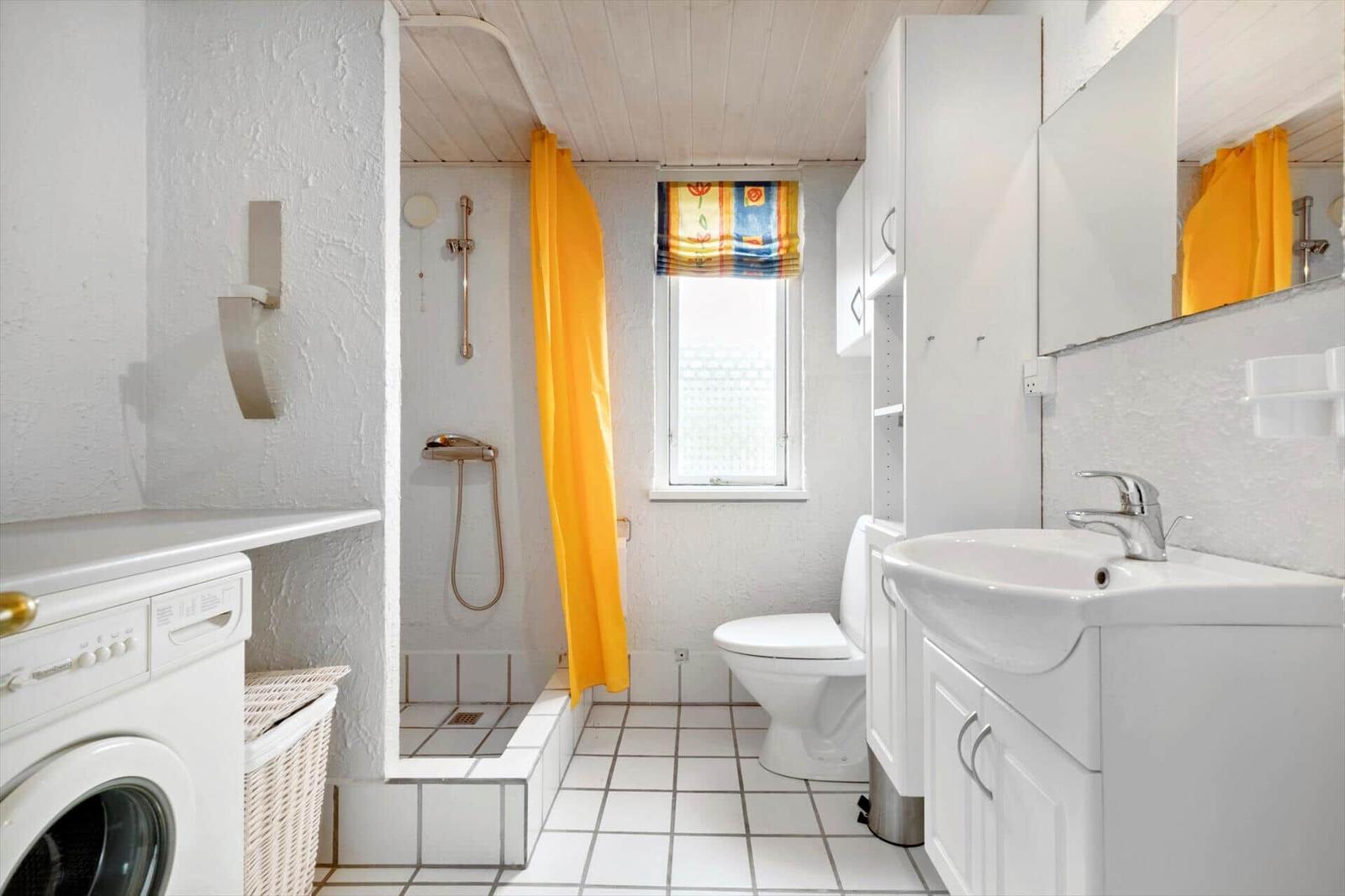 Bathroom with shower, toilet, and washing machine. Yellow shower curtain and window with roller blind.